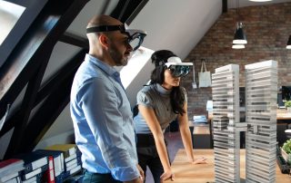 Due professionisti, un uomo e una donna, indossano visori per realtà aumentata e osservano il modello 3D olografico di un edificio che fluttua sopra un tavolo in un ufficio moderno.