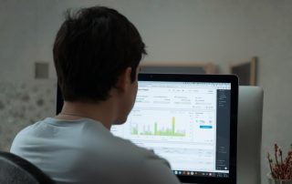 Inquadratura da dietro le spalle di un giovane intento a studiare un report su un monitor di un computer.