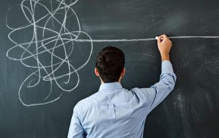 Uomo in camicia visto di spalle mentre traccia con il gesso una linea bianca retta e decisa su una lavagna, facendola emergere da un ammasso confuso di linee e scarabocchi.