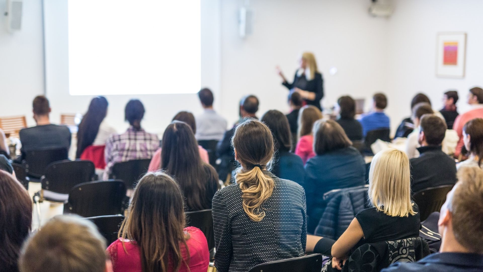 Persone di spalle sedute in un'aula che ascoltano una relatrice