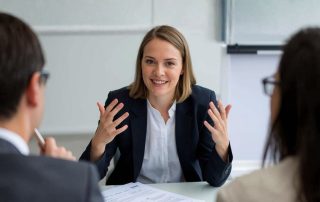 Primo piano di una giovane donna che gesticola con enfasi per spiegare un concetto a due interlocutori, di cui si intravedono solo le teste in primo piano.