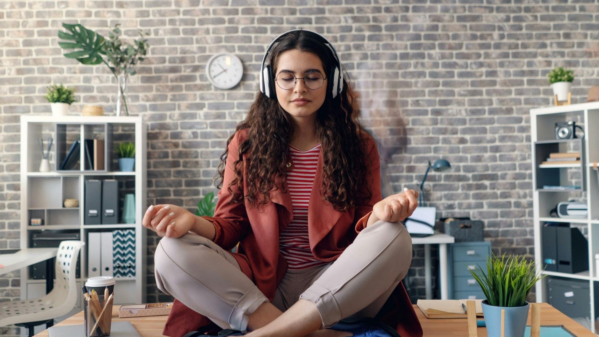 Una giovane donna seduta sulla scrivania di un ufficio moderno in posizione di meditazione (loto). Indossa delle cuffie, ha gli occhi chiusi e un'espressione serena, circondata da un ambiente di lavoro luminoso.