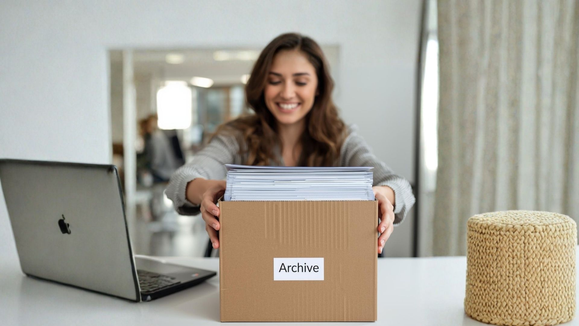 Una ragazza in un ufficio luminoso ripone vecchi documenti in uno scatolone con la scritta 'Archivio', mostrando un'espressione felice.
